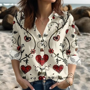 Könnte beinhalten: Cremefarbenes Button-Down-Shirt mit Blumen- und Herzmuster. Das langärmlige Hemd hat einen V-Ausschnitt und ist mit roten Herzen, Blumen und dunkelgrünen Ranken verziert. Die Manschetten haben ein Blumenmuster.