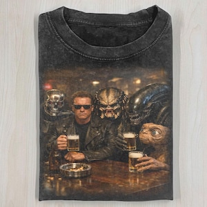 May include: Dark gray t-shirt featuring a humorous graphic of Arnold Schwarzenegger and other movie characters at a bar, each holding a beer. The image is printed on a folded, crew neck tee.