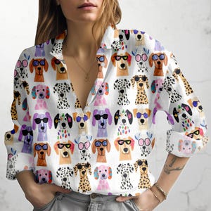Könnte beinhalten: Weißes Button-Down-Hemd mit einem sich wiederholenden Muster von Cartoon-Hunden mit Sonnenbrillen. Die Hunde sind in verschiedenen Farben und Rassen, darunter Dalmatiner. Das Hemd hat lange Ärmel und einen Kragen.