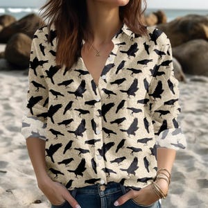 Könnte beinhalten: Beige Knopfleiste mit schwarzem Krähenmuster. Das langärmlige Hemd hat einen V-Ausschnitt und ist aus leichtem Stoff gefertigt. Das Krähendesign wiederholt sich auf dem gesamten Hemd.