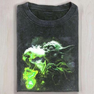 Könnte beinhalten: Dunkelgraues T-Shirt mit einer Grafik von Yoda, der grünen Rauch ausatmet. Das Bild zeigt Yoda in einem grünen Gewand mit großen Ohren, der einen grünen Gegenstand hält. Der Rauch und der Gegenstand leuchten neongrün.