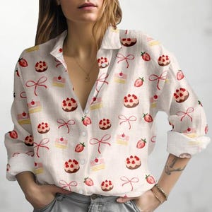 Può includere: Camicia color crema con bottoni con un motivo ripetuto di fragole, torte e fiocchi rossi. La camicia a maniche lunghe ha un colletto classico ed è realizzata in tessuto leggero.