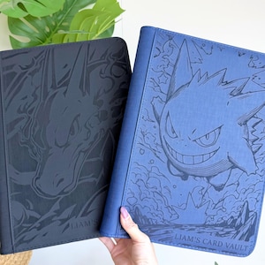 Puede incluir: Dos carpetas rectangulares para tarjetas con cubiertas texturizadas. La carpeta negra presenta una ilustraci&oacute;n oscura de Charizard, mientras que la azul muestra una ilustraci&oacute;n de Gengar y el texto "LIAM'S CARD VAULT" en la parte inferior.