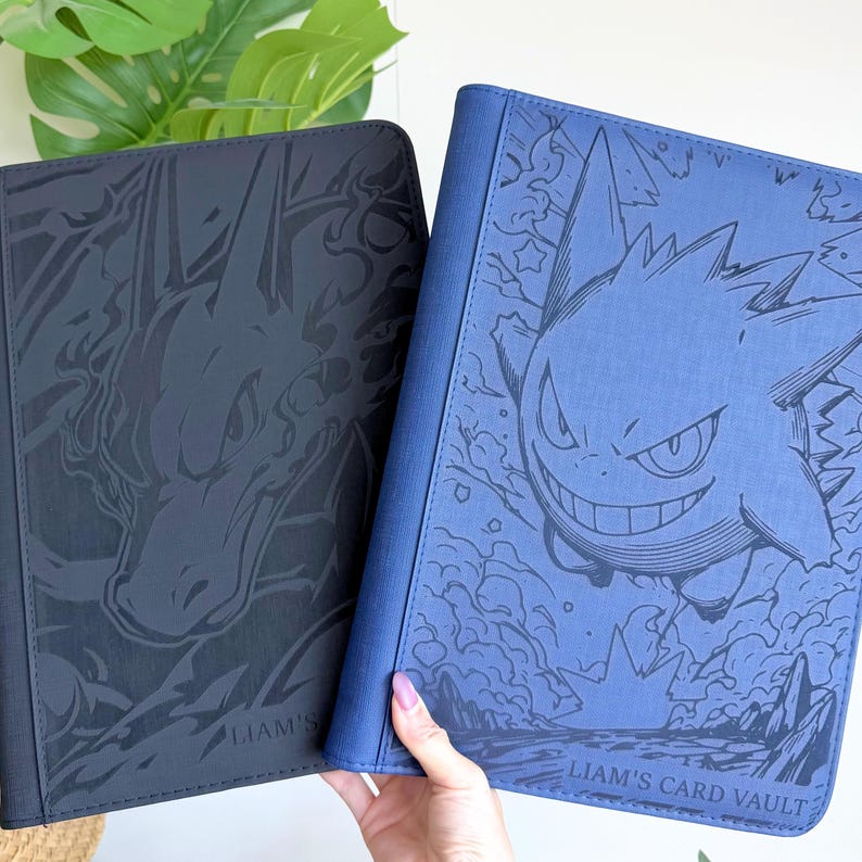 Puede incluir: Dos carpetas rectangulares para tarjetas con cubiertas texturizadas. La carpeta negra presenta una ilustraci&oacute;n oscura de Charizard, mientras que la azul muestra una ilustraci&oacute;n de Gengar y el texto "LIAM'S CARD VAULT" en la parte inferior.