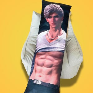 May include: A body pillow featuring a digitally rendered male figure with silver hair, wearing a white V-neck shirt and a black belt. The pillow is rectangular and rests on a light gray beanbag chair against a yellow background.