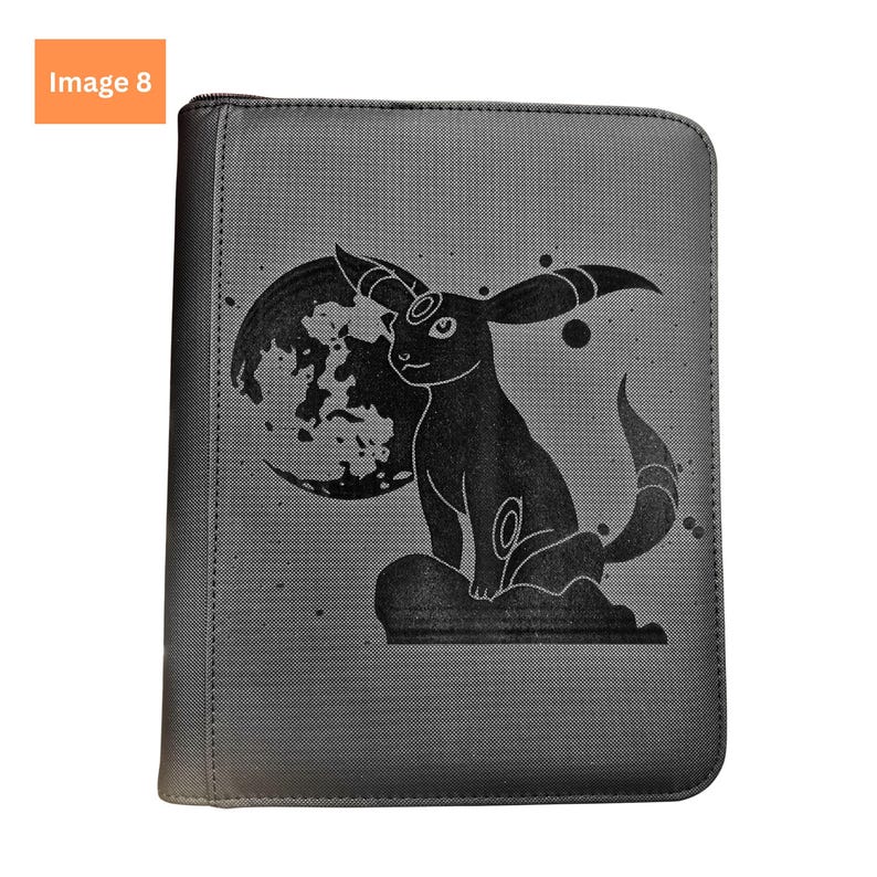 Puede incluir: Una carpeta rectangular negra con cremallera que presenta una ilustraci&oacute;n gr&aacute;fica oscura de un personaje Umbreon sentado frente a una luna grande. El dise&ntilde;o incluye peque&ntilde;os acentos circulares y una superficie texturizada en la cubierta.