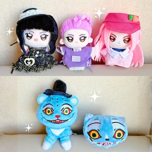 May include: A selection of handmade plush dolls with detailed faces and diverse hairstyles. The dolls are dressed in various outfits, including a black dress, a purple top, and a pink cap. A blue cat plush with a top hat and a smiling face is also featured.