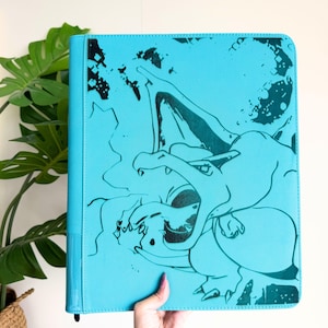 Puede incluir: Una carpeta de color cian brillante con una ilustración gráfica en verde azulado oscuro de la criatura Charizard en la portada. El diseño muestra a la criatura con las alas extendidas y la boca abierta, sobre un fondo de formas abstractas.