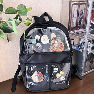 Puede incluir: Mochila negra con bolsillos frontales transparentes que muestran coloridos pines esmaltados. La mochila tiene un asa superior, correas ajustables y un bolsillo frontal. Los pines presentan varios personajes de estilo anime en colores vibrantes.
