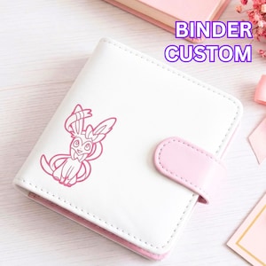 May include: A white square binder with a pink snap closure and a pink line art illustration of the Sylveon character on the front cover. The text BINDER CUSTOM is printed in purple in the upper right corner of the image.
