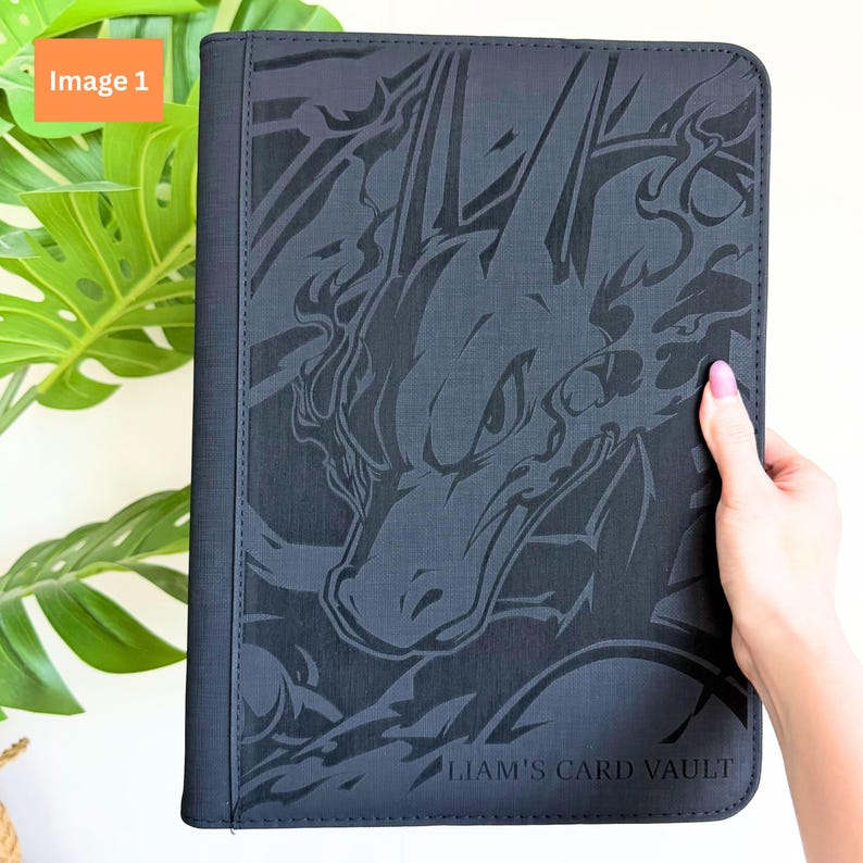 Puede incluir: Una carpeta de tarjetas rectangular de color negro con un gr&aacute;fico oscuro en relieve de una criatura similar a un drag&oacute;n en la portada. La esquina inferior derecha incluye el texto "LIAM'S CARD VAULT" impreso en una fuente sans-serif.