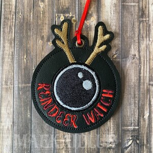 May include: A black felt ornament with a reindeer face and the text "Reindeer Watch" in red embroidery. The reindeer has gold antlers and a white eye.