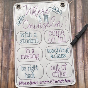 May include: White sign with the text "Where is the Counselor?" and a list of reasons why the counselor may be unavailable. The text is in a purple and teal font. The sign is hanging on a wooden surface.