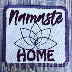 May include: Embroidered patch with a light blue background and a purple border. The patch features the word "Namaste" in cursive above a lotus flower design and the word "HOME" below. The patch is square with rounded corners.