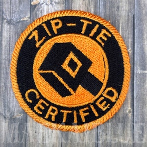 May include: A round, orange and black embroidered patch with the text "Zip-Tie Certified" in a circle around a stylized black logo.