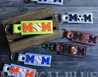 May include: Six keychains with the word "MOM" embroidered on them. Each keychain features a different sport ball: baseball, soccer, volleyball, basketball, and football. The keychains are made of different materials, including felt, leather, and fabric.