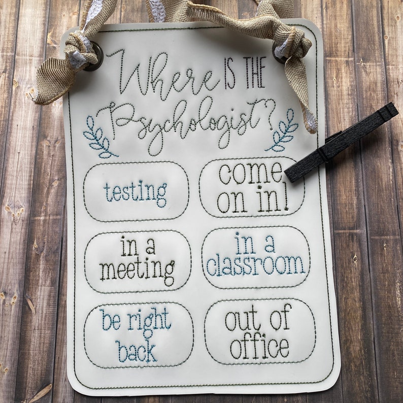 May include: A white sign with black and blue embroidery that reads "Where is the Psychologist?" with a series of options below: "testing", "come on in!", "in a meeting", "in a classroom", "be right back", "out of office".