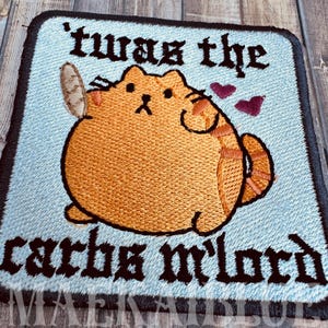 May include: Embroidered patch featuring a cartoon cat holding a baguette, with the text "'twas the carbs m'lord" in gothic lettering. The cat is orange with brown stripes, set against a light blue background with a dark blue border. Two purple hearts are also present.