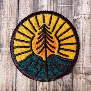 May include: A round embroidered patch with a yellow sun, orange tree, and teal green mountains. The sun has black rays and the tree has a black outline.