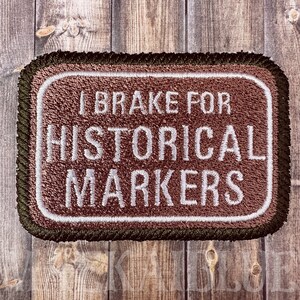 May include: Embroidered patch with the text "I Brake for Historical Markers" in white on a brown background. The patch has a white border and a dark green and brown rope-like edge.