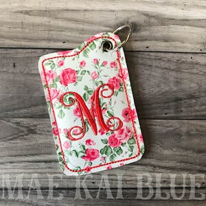 May include: A white fabric key chain with a red floral pattern and a red embroidered letter "M". The key chain has a silver metal ring.