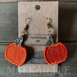 May include: A pair of orange pumpkin-shaped earrings with green stems and gold hooks. The earrings are handmade by Mae Kai Blue.