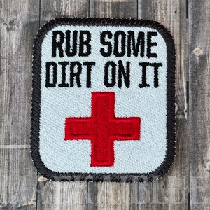 May include: Embroidered patch with the text "RUB SOME DIRT ON IT" above a red cross symbol. The patch has a light blue background, black border, and is likely for a first aid kit or medical theme.