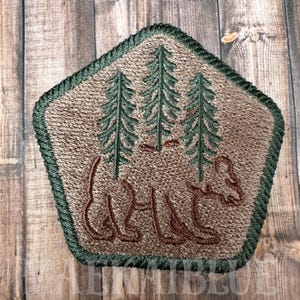 Peut inclure: Patch brodé avec une silhouette d'ours brun et trois sapins verts. Le patch est en forme de pentagone avec une bordure verte.