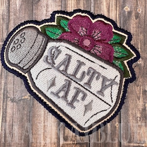 May include: Embroidered patch of a salt shaker with a flower on top. The salt shaker is white with the words "Salty AF" on it. The flower is pink with green leaves.