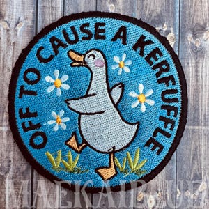 May include: Embroidered patch with a blue background and a black border. The patch features a cartoon duck with the text "Off to Cause a Kerfuffle" in black. White daisies with yellow centers are scattered around the duck.