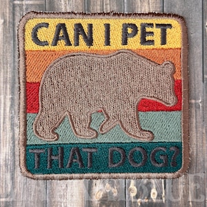 May include: A brown embroidered patch with a retro-style design. It features a brown bear silhouette with a sunset background and the text "CAN I PET THAT DOG?"