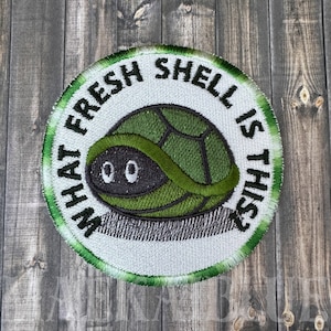 May include: A white, round embroidered patch with a green border. The patch features a cartoon turtle with the text "What Fresh Shell Is This?"
