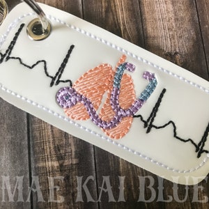 May include: White key chain with a pink and purple embroidered stethoscope and lungs with a black EKG line. The key chain has a silver key ring.
