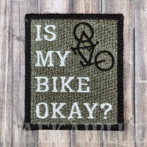 Peut inclure: Un patch brodé vert avec des coutures noires et du texte blanc qui dit "Is my bike okay?" avec un graphique de vélo stylisé.