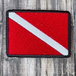 May include: A red and white scuba diving flag patch with a black border. The flag is made of a textured fabric.