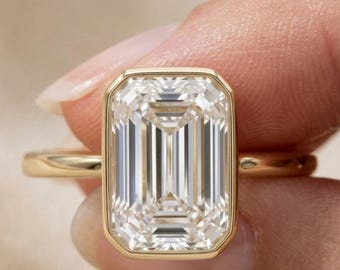 May include: A gold ring featuring a large, rectangular emerald-cut clear gemstone set in a smooth bezel setting. The band is thin and polished gold. The logo DK JEWELS is visible in the upper left corner of the image.