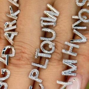 Könnte beinhalten: Nahaufnahme von silberfarbenem Schmuck, der Wörter buchstabiert. Die Buchstaben sind mit kleinen, funkelnden Steinen besetzt. Der Schmuck wird auf hellem Hautton-Hintergrund präsentiert.