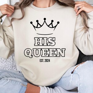 May include: A cream-coloured sweatshirt with a black crown and the text "HIS QUEEN EST. 2024".