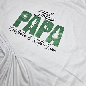 Op de afbeelding: Wit T-shirt met een groene afbeelding met de tekst "Stolzer Papa", een zon, een visser en een boom. De tekst "Konstantin & Kate-Linn" staat eronder in een scriptlettertype.