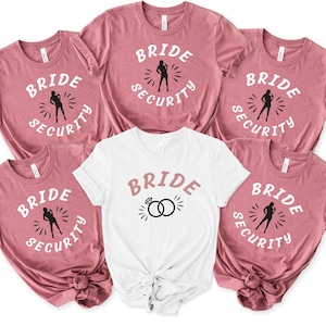 以下が含まれることがあります： 白地に「Bride Security」と書いてあるピンクのTシャツが5枚。1枚は、警備員のグラフィックの代わりにリングのデザインが施されています。