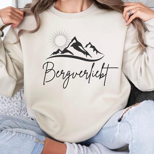 Könnte beinhalten: Ein cremefarbener Pullover mit einem schwarzen Bergpanorama-Motiv und dem Schriftzug "Bergverliebt" auf der Vorderseite.