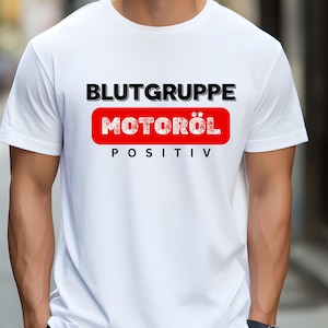 Puede incluir: Camiseta blanca con el texto "BLUTGRUPPE MOTORÖL POSITIV" en negro y rojo. El texto está centrado en el pecho. La camiseta es de manga corta y parece estar hecha de un material suave.
