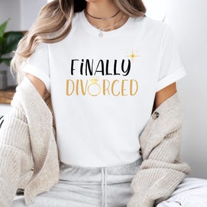 Puede incluir: Camiseta blanca con texto negro que dice "FINALMENTE DIVORCIADA" con un anillo dorado y un gráfico de brillo.