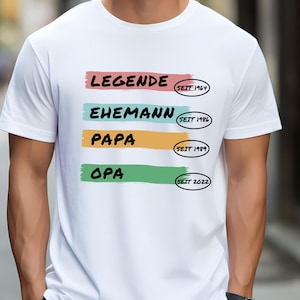 May include: White t-shirt with a colourful graphic that reads "Legende seit 1964", "Ehemann seit 1986", "Papa seit 1989", and "Opa seit 2022".