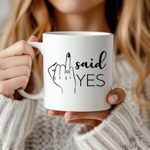 Könnte beinhalten: Weiße Keramiktasse mit einer schwarzen Illustration einer Hand, die den Mittelfinger zeigt, mit einem Ring am Ringfinger. Der Text "said YES" steht unter der Hand.