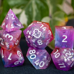 May include: A set of seven polyhedral dice in shades of purple, pink, and blue. The dice have white numbering and are embedded with glitter and star-shaped accents. One die features a ghost design.