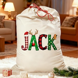 May include: A large, off-white Christmas gift bag with a red drawstring closure. The word "JACK" is printed on the front in festive colors and patterns, including a Santa hat, antlers, and a gingerbread man. The bag is sitting on a white rug.
