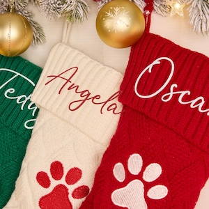 May include: Three knitted Christmas stockings in green, cream, and red, each with a name embroidered on the cuff. The green stocking has a fishbone design, while the cream and red stockings feature paw prints. Gold ornaments and festive greenery complete the holiday scene.