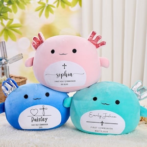 Puede incluir: Tres peluches con forma de ajolote en rosa, azul y turquesa. Cada uno tiene una cara sonriente y una etiqueta blanca con un nombre, una cruz y una fecha, lo que indica un tema de Primera Comunión. El rosa dice "Sophia", el azul dice "Paisley" y el turquesa dice "Evenly Joshua".