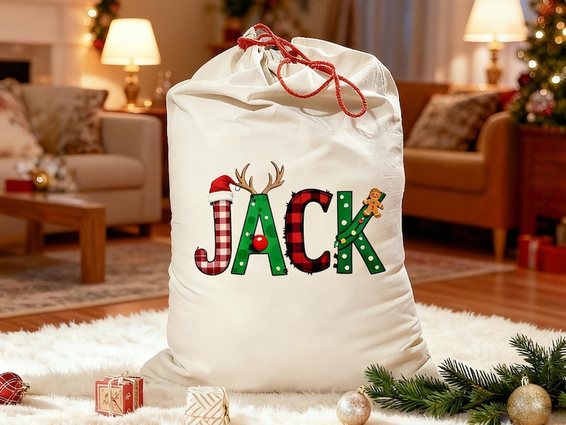 May include: A large, off-white Christmas gift bag with a red drawstring closure. The word "JACK" is printed on the front in festive colors and patterns, including a Santa hat, antlers, and a gingerbread man. The bag is sitting on a white rug.